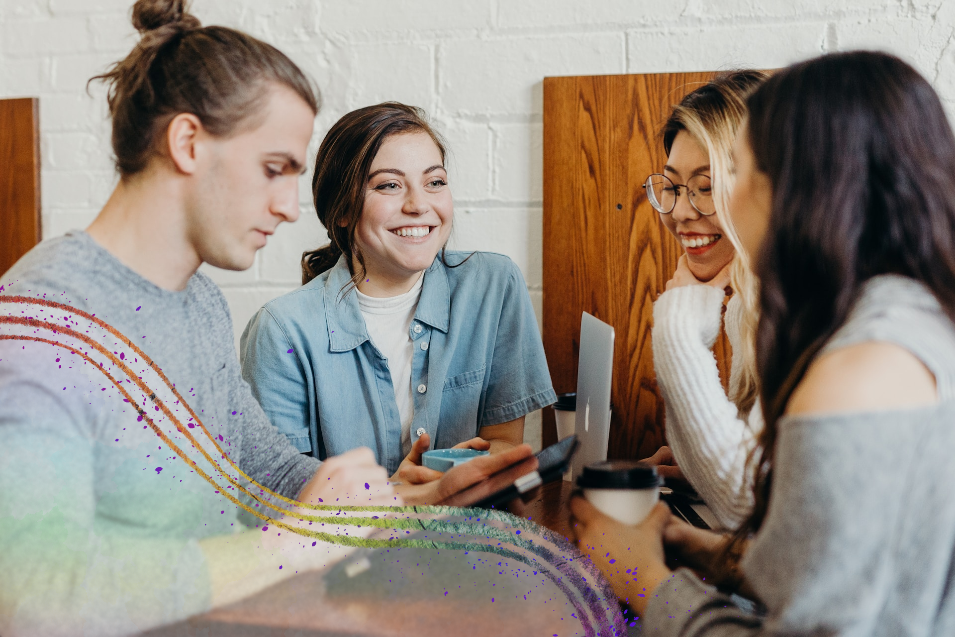 A group of young people enjoying eachothers company (except the guy who is busy doom-scrolling on his phone) around a table with a brick wall in the background and a wooden decoration behind the smiling girl with the glasses